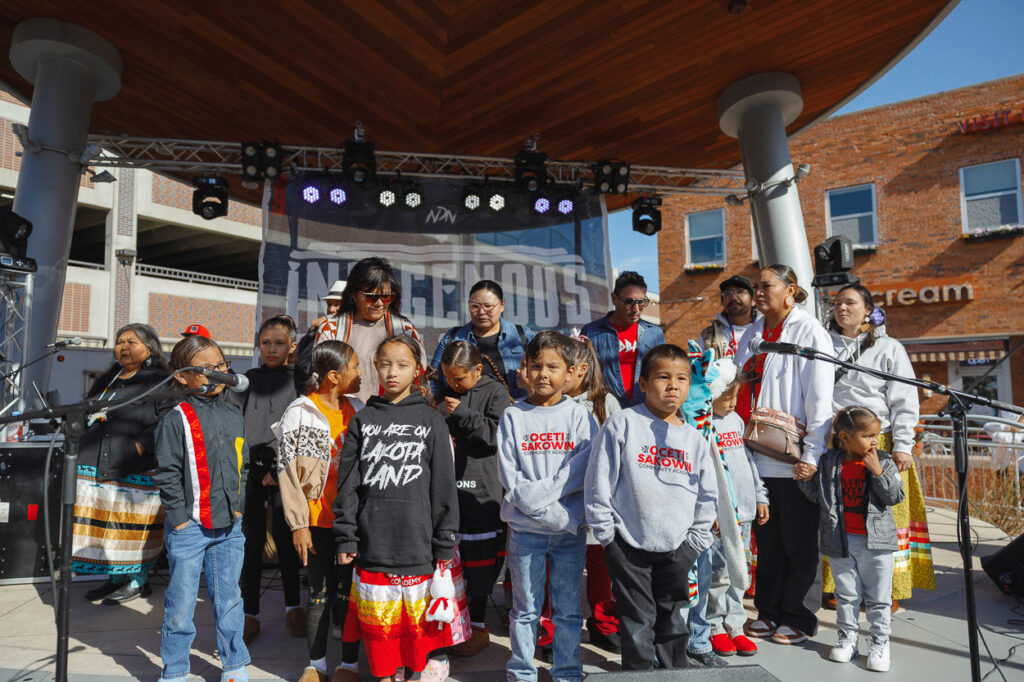 A group of about a dozen children stand on a stage in front of microphones. Six adults are standing behind them. 