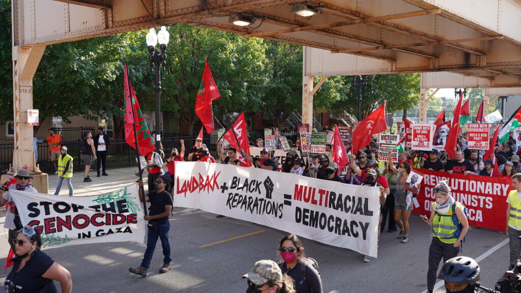 Photo description: Indigenous Peoples' at the March on DNC display a Landback + Black Reparations = Multiracial Democracy banner.   